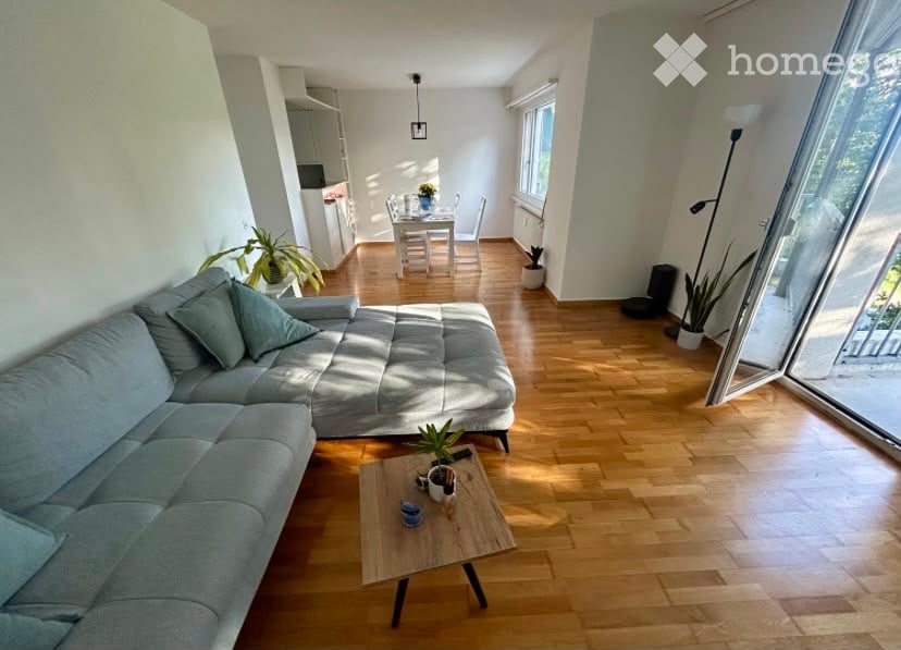 Bright living room with light grey sectional sofa, wooden coffee table, dining area with white table and chairs, and several potted plants. Natural light streams in from large windows and a glass door leading to a balcony.