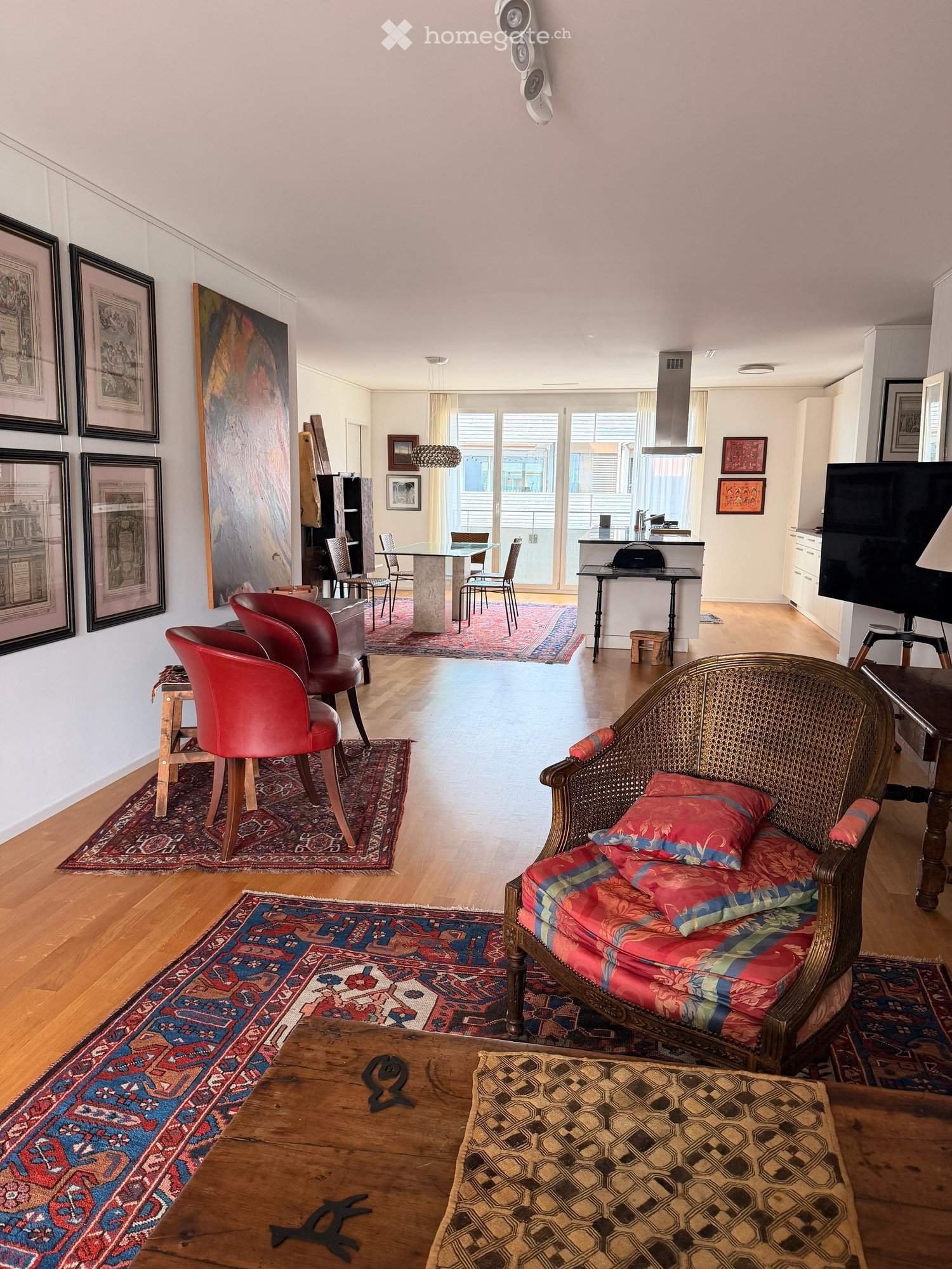 Open-plan living and dining area with wooden flooring, colorful rugs, a red leather sofa, a wicker armchair, a glass dining table, and a grand piano.