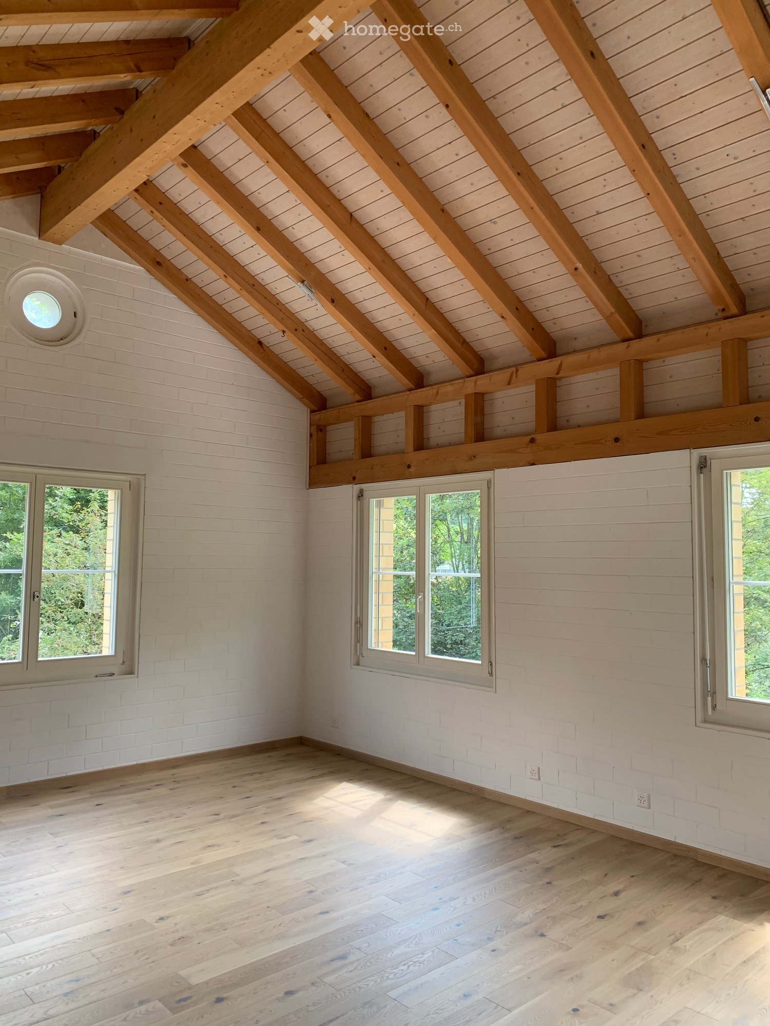 Bright, airy attic space with exposed wooden beams, white shiplap walls, and large windows allowing natural light to flood the room. The floor is made of light-colored hardwood planks.