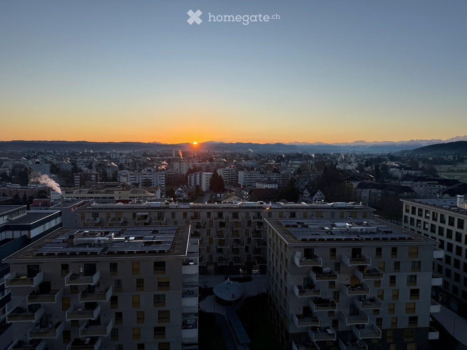 A modern apartment complex with multiple buildings, balconies, and solar panels on the rooftops, set against a cityscape with a sunset view of mountains in the background.