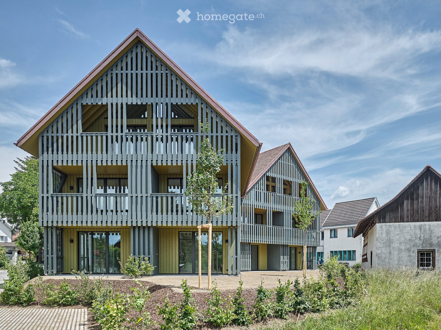 Modern two-story house with a distinctive wooden facade, large windows, and a raised ground floor. The house features a triangular roof and is surrounded by greenery and a paved pathway.