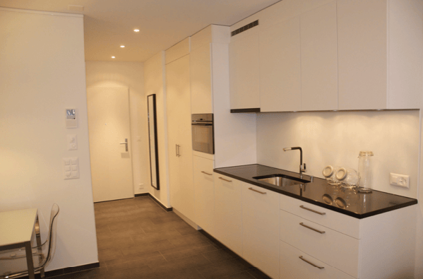 Kitchen. Modern kitchen with white cabinets, black countertop, stainless steel sink, and a small oven. The floor is dark wood, and there is a glass bottle on the counter.