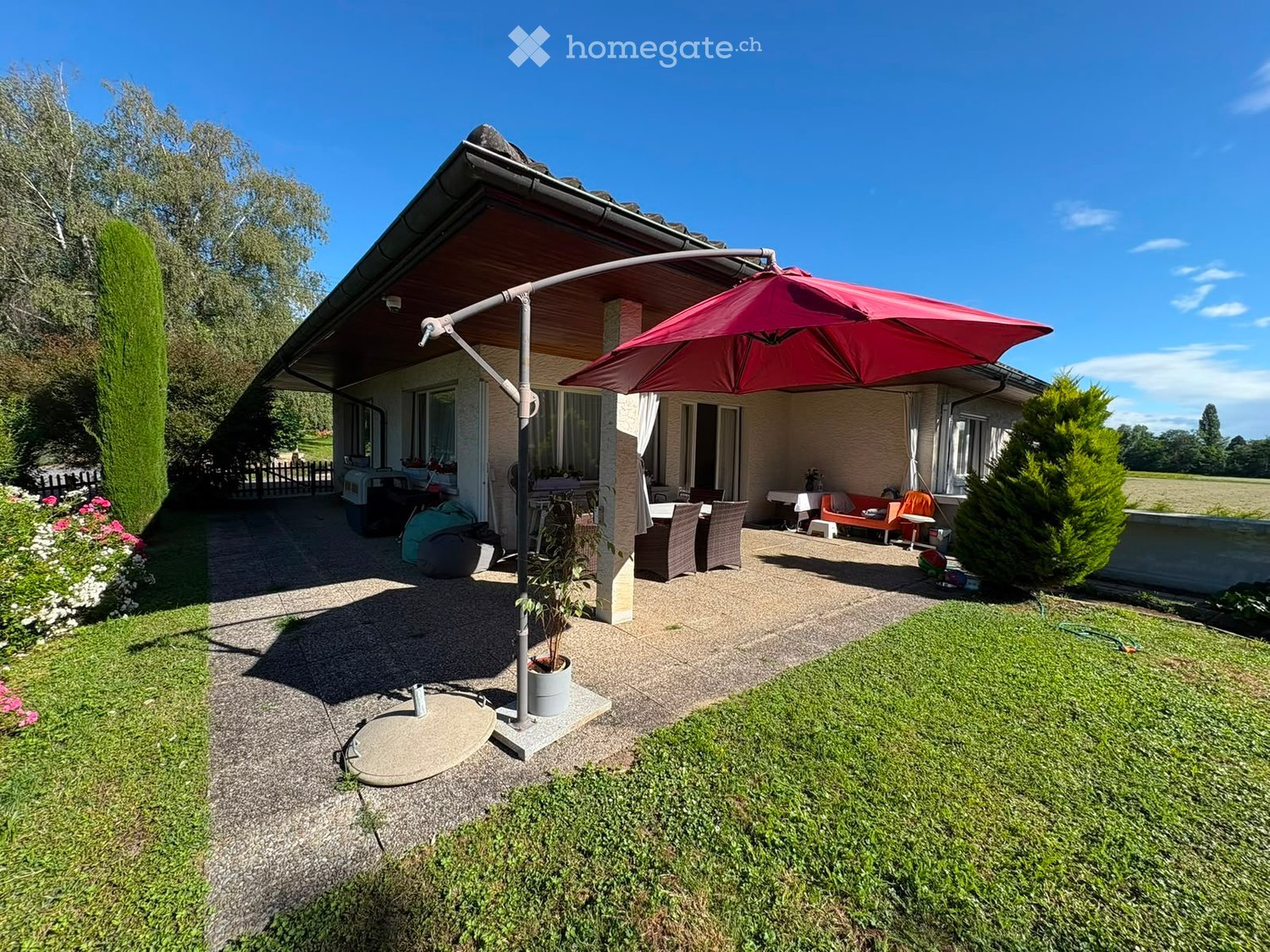 Outdoor patio area with a large umbrella, dining table, chairs, and a small garden with grass and flowers.