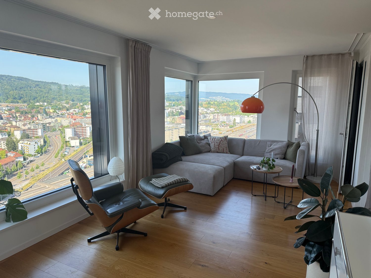 Modern living room with large windows offering a scenic city view, featuring a grey sectional sofa, a leather armchair, a small round table, a floor lamp, and a potted plant.