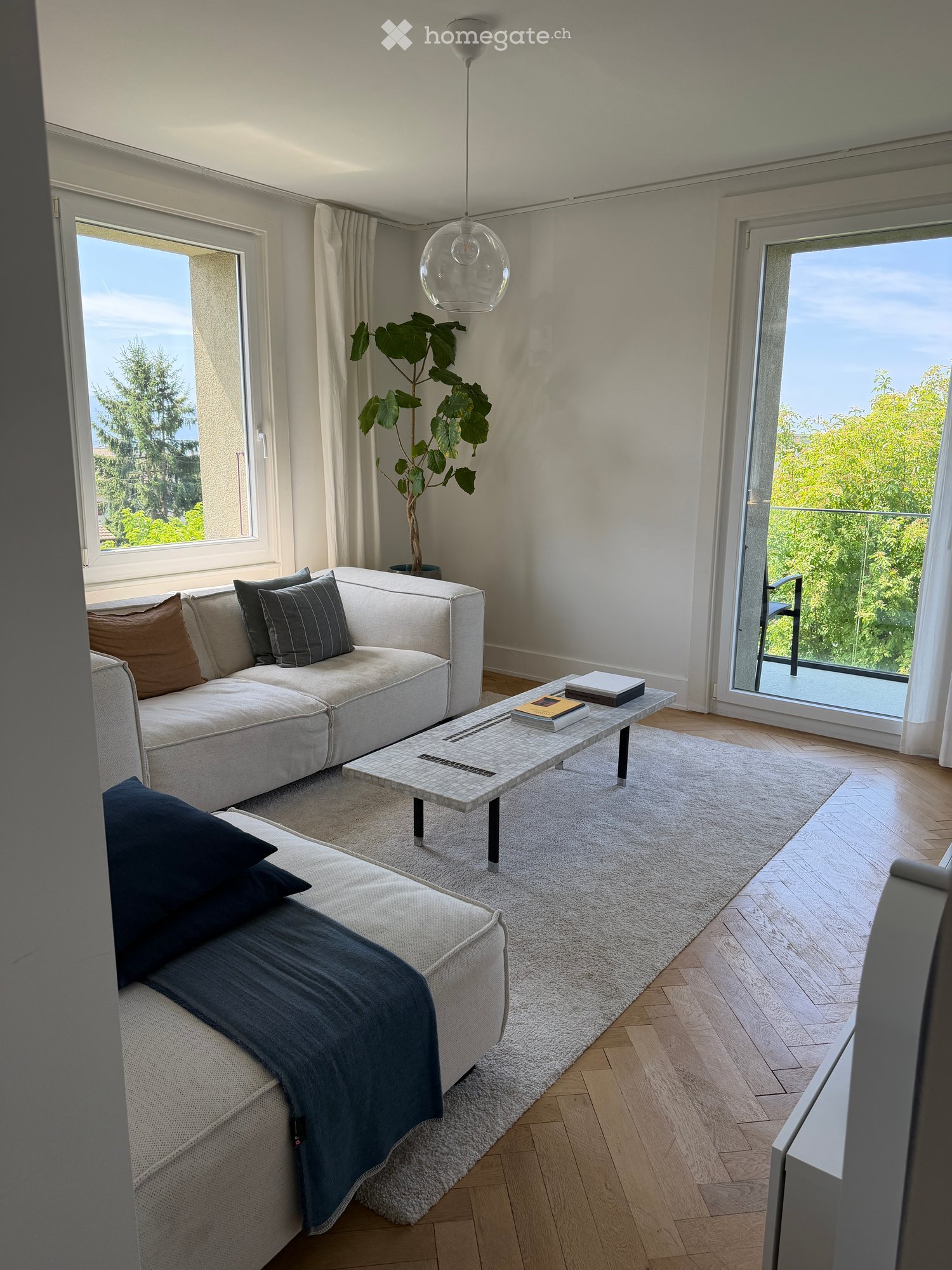 Modern living room with a white sectional sofa, coffee table, books, a potted plant, and a hanging lamp. The room features large windows and a sliding glass door leading to a balcony.
