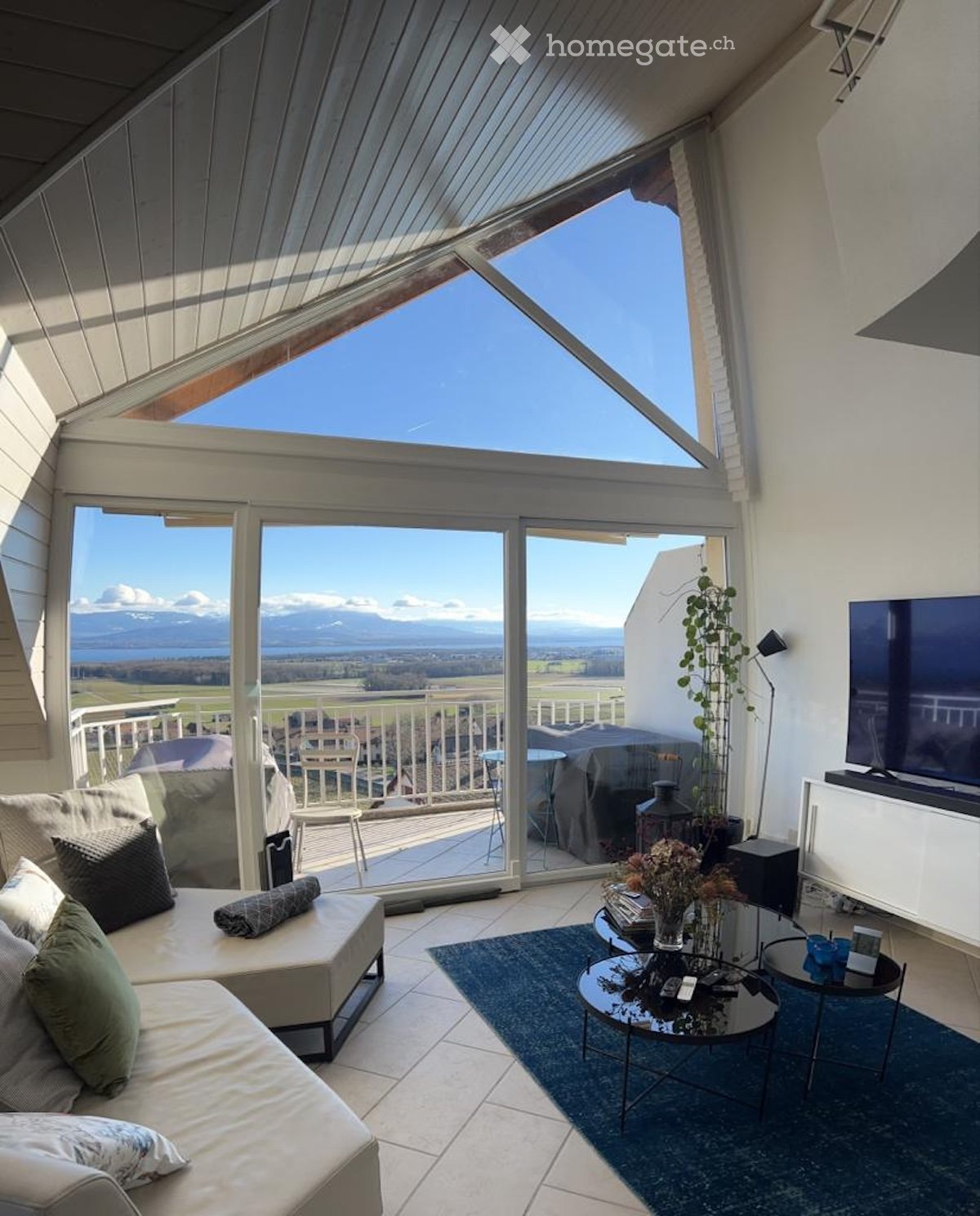 Bright living room with large windows and a view of the mountains, featuring a sofa, coffee table, TV, and a small balcony area with outdoor furniture.