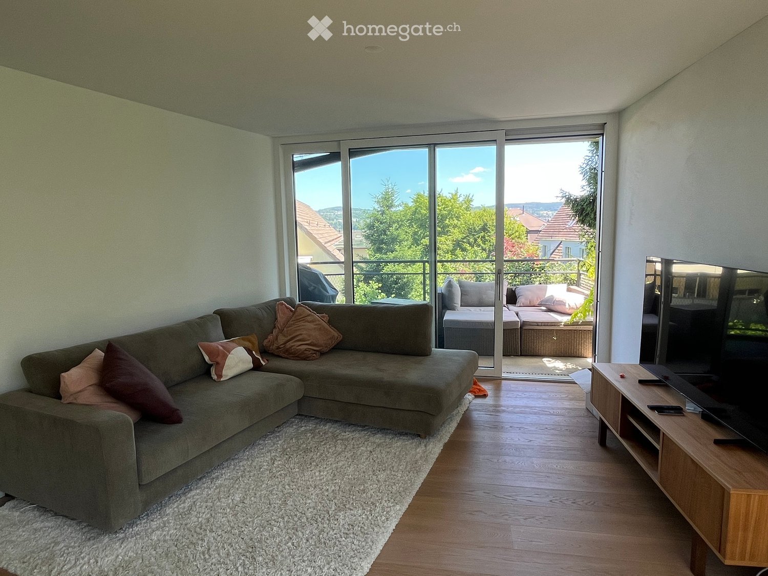 Modern living room with a grey sectional sofa, multiple throw pillows, a flat-screen TV on a wooden stand, and a view of greenery through large glass doors leading to a balcony.