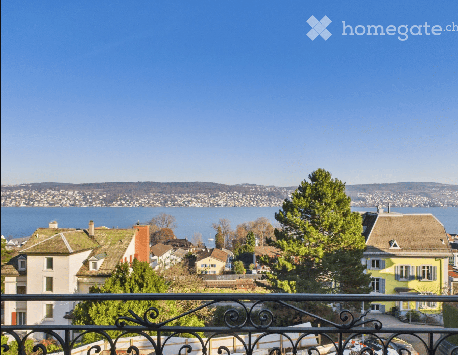 Balcony overlooking a scenic lakeside town with houses, trees, and distant water.