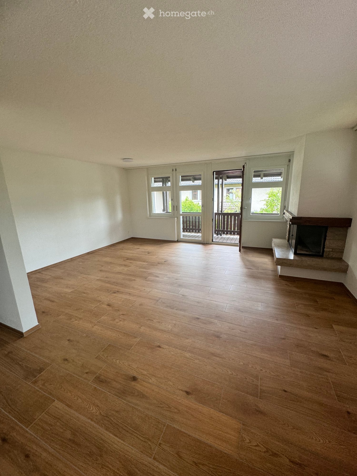 Empty room with wooden flooring, white walls, and a fireplace on the right side. There are large windows and a door leading to a balcony.