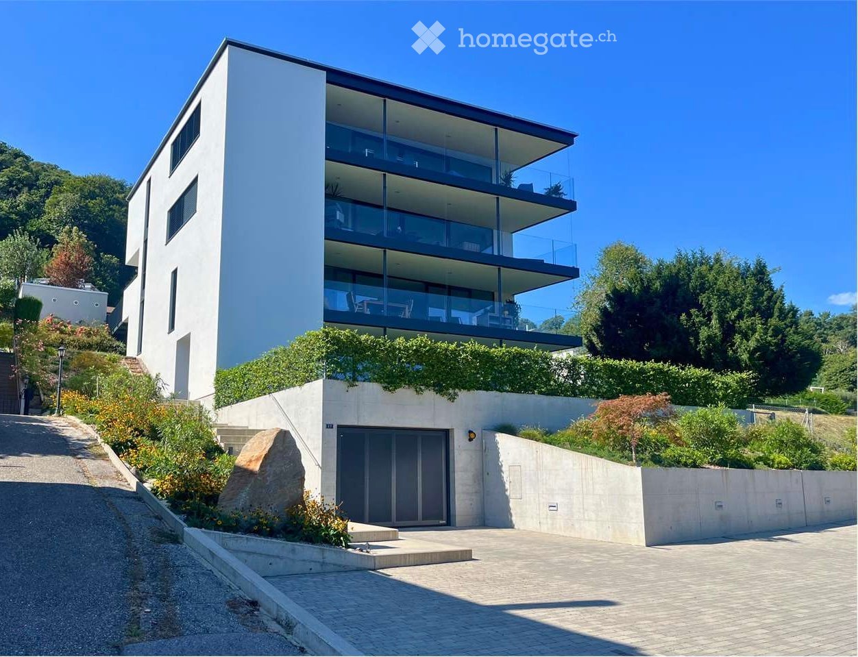 Modern two-story house with white facade, large glass balcony, garage, and landscaped garden.