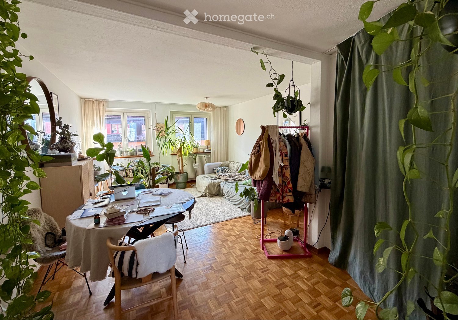 A cozy, multi-functional room with a wooden floor, large windows, a sofa, a work table with a laptop, and several potted plants. The room features a coat rack with jackets and a mirror on the wall.