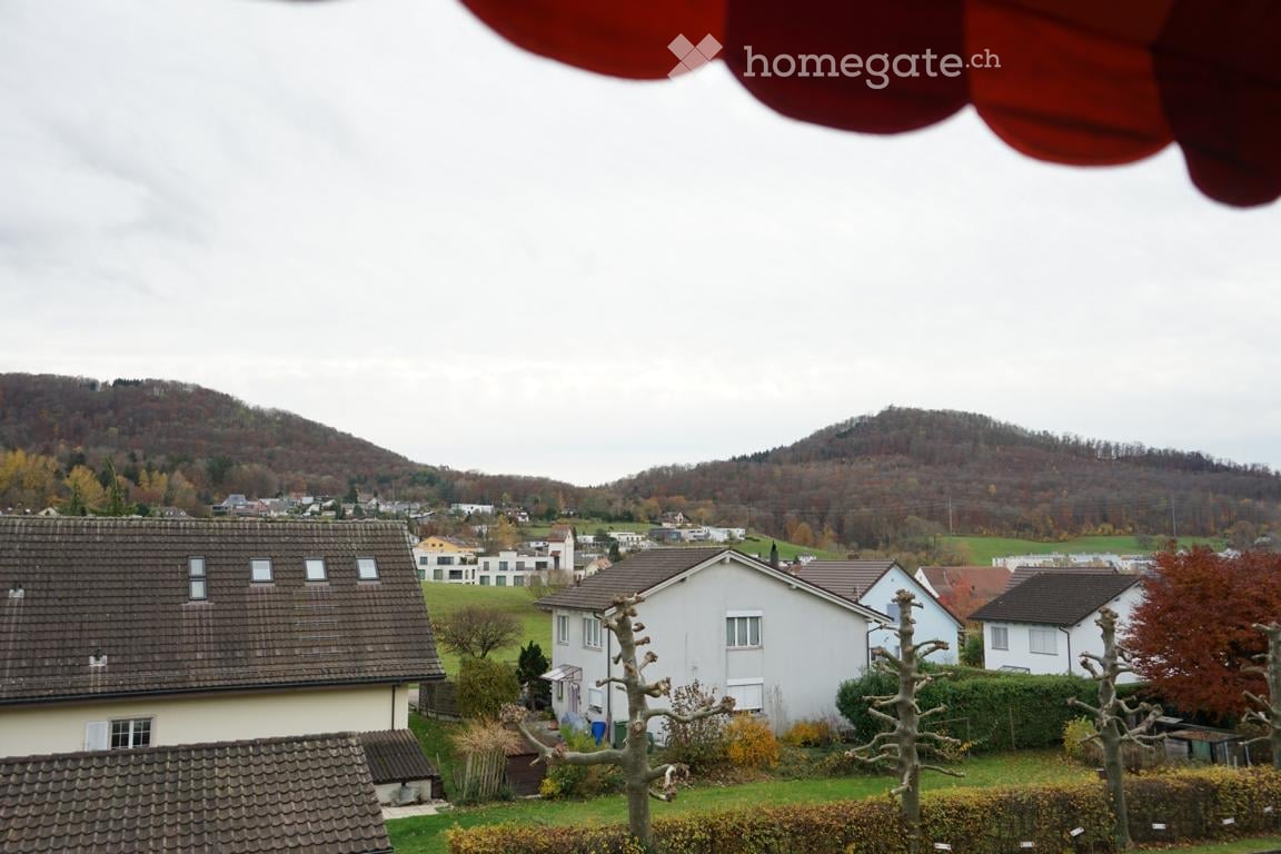 Residential area with multiple houses, surrounded by green fields and hills under a cloudy sky.