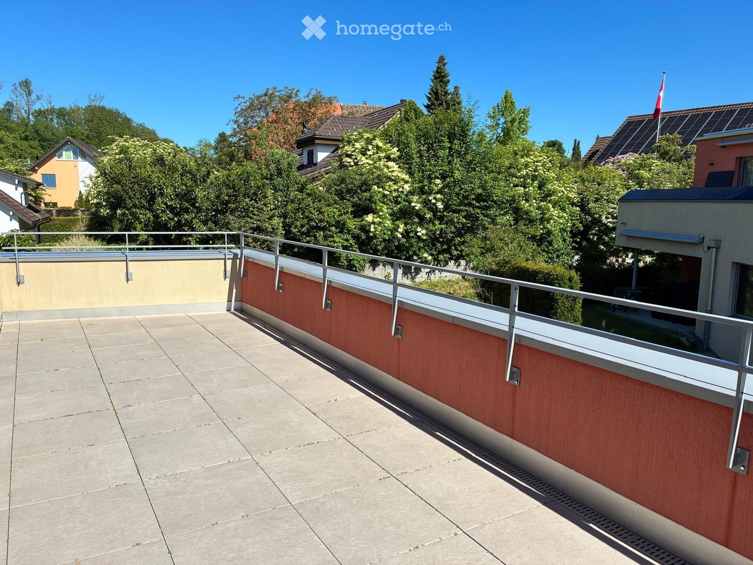 Roof terrace with concrete tiles, metal railing, and red wooden privacy wall. Surrounded by trees and neighboring houses.