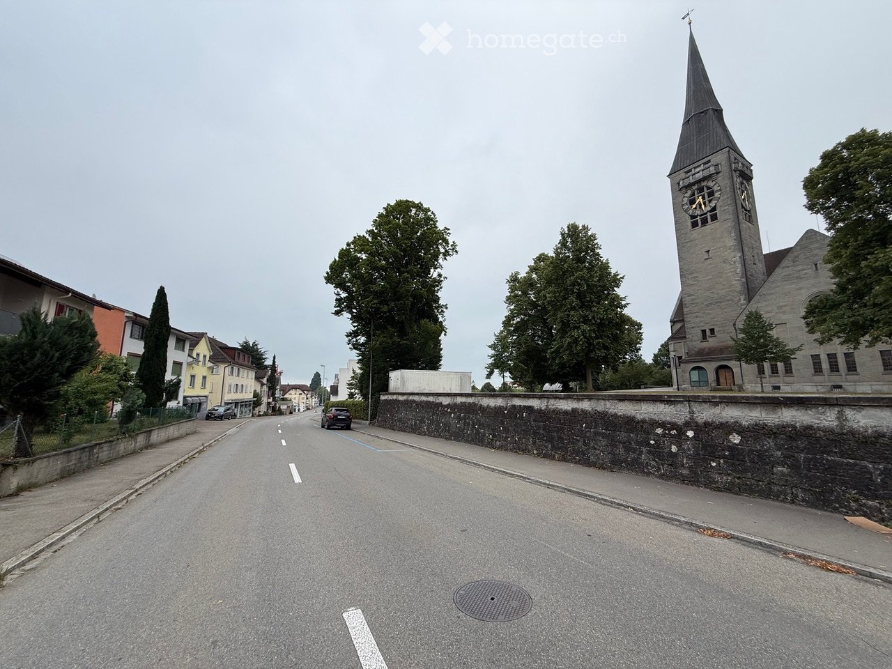 A quiet street lined with buildings, including a prominent church with a tall steeple and clock tower, surrounded by trees and parked cars.