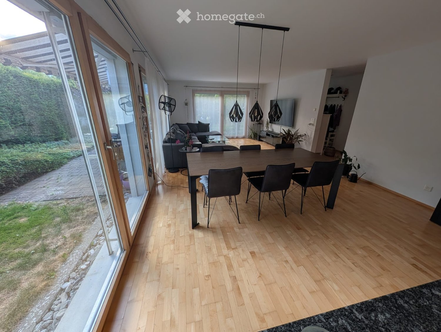Bright dining area with wooden flooring, large glass doors leading to outdoor space, dining table with chairs, sofa in adjacent living area, fans, and pendant lights.