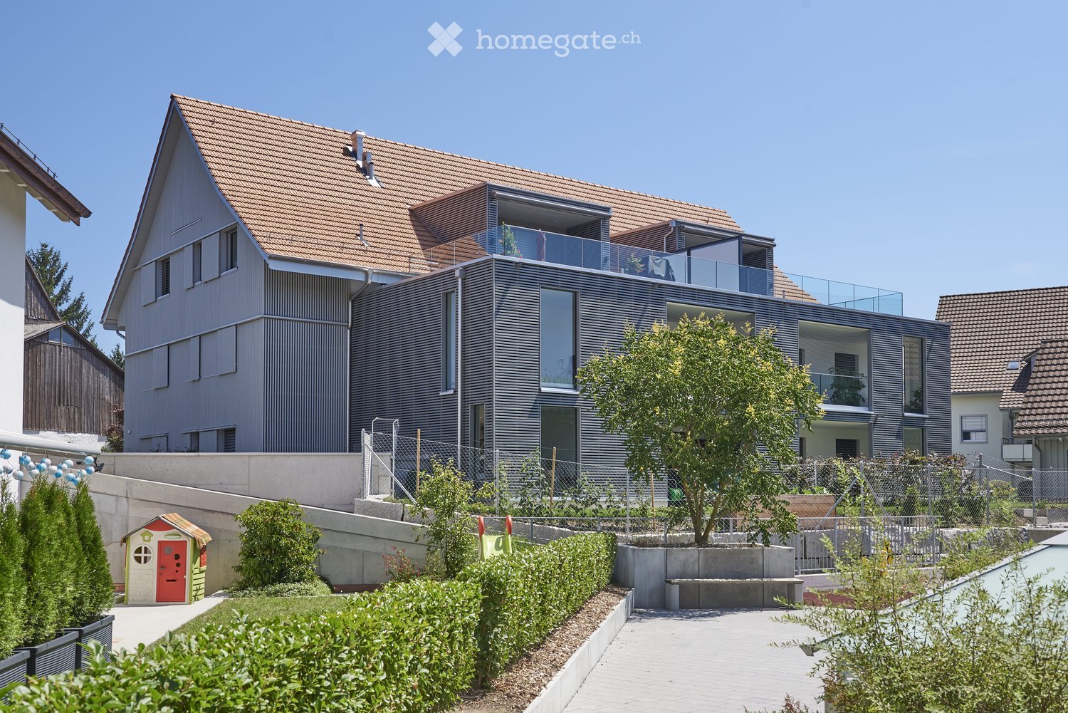 Modern two-story house with a combination of grey and brown exterior, featuring large windows, a balcony, and a well-maintained garden with a small playhouse.