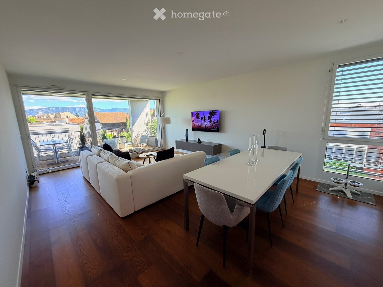 Modern living and dining area with a white sectional sofa, a white dining table with chairs, a television mounted on the wall, and a view of the mountains through large windows and a balcony.