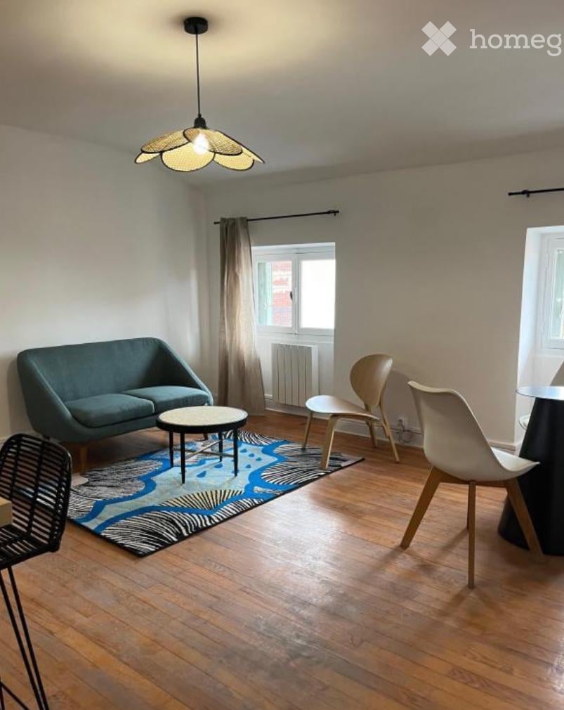 Modern living room with wooden flooring, a blue sofa, a coffee table, and several chairs arranged around a patterned rug.