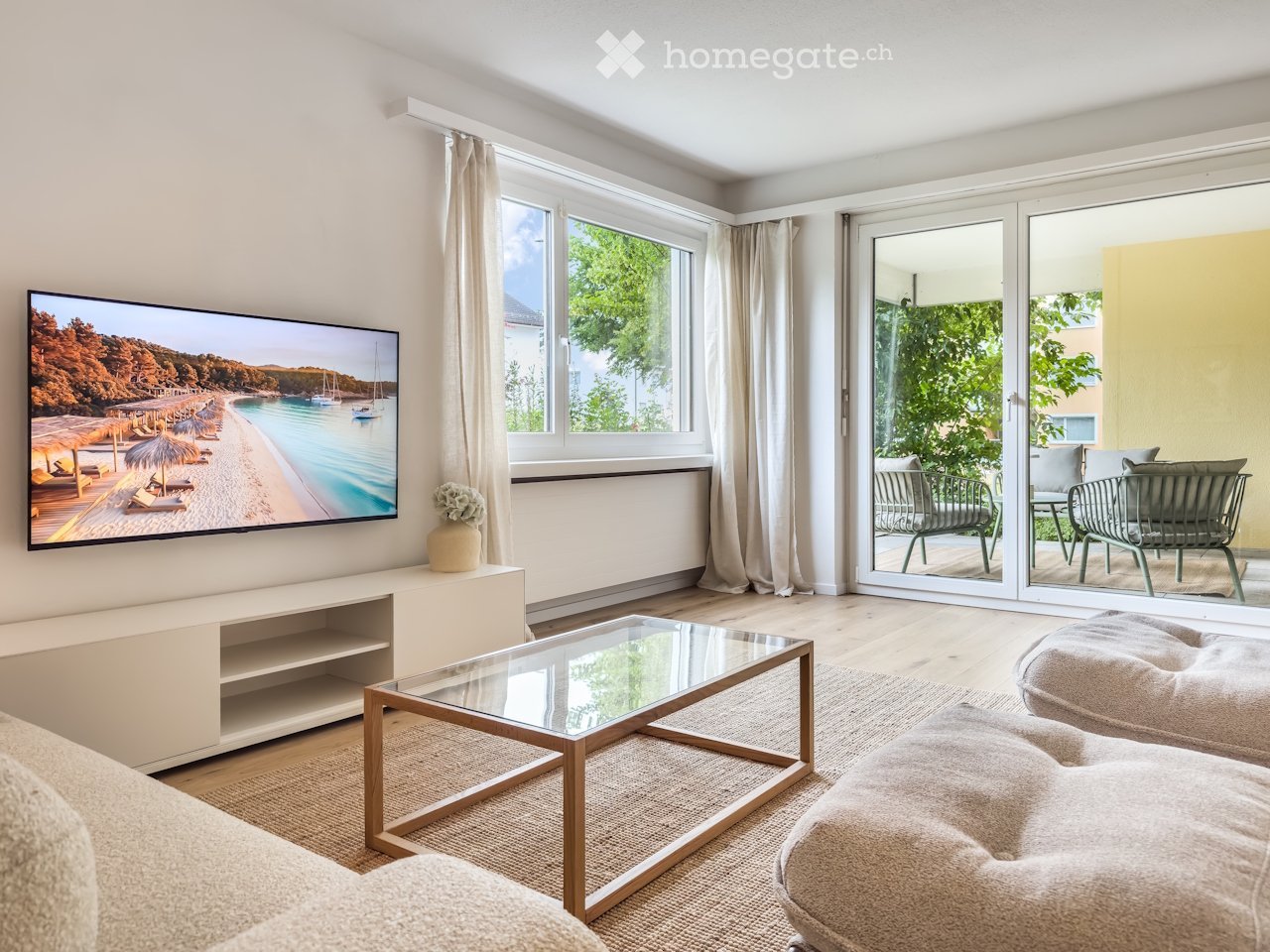 Modern living room with large windows, a flat-screen TV, a glass coffee table, a sofa, and a view of a balcony with outdoor seating.