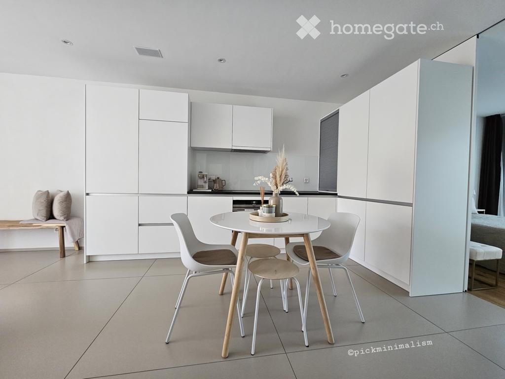 Modern, minimalist dining area with a white dining table, four chairs, a bench, and a small vase with dried flowers on the table. The room features white cabinets and a neutral color palette.
