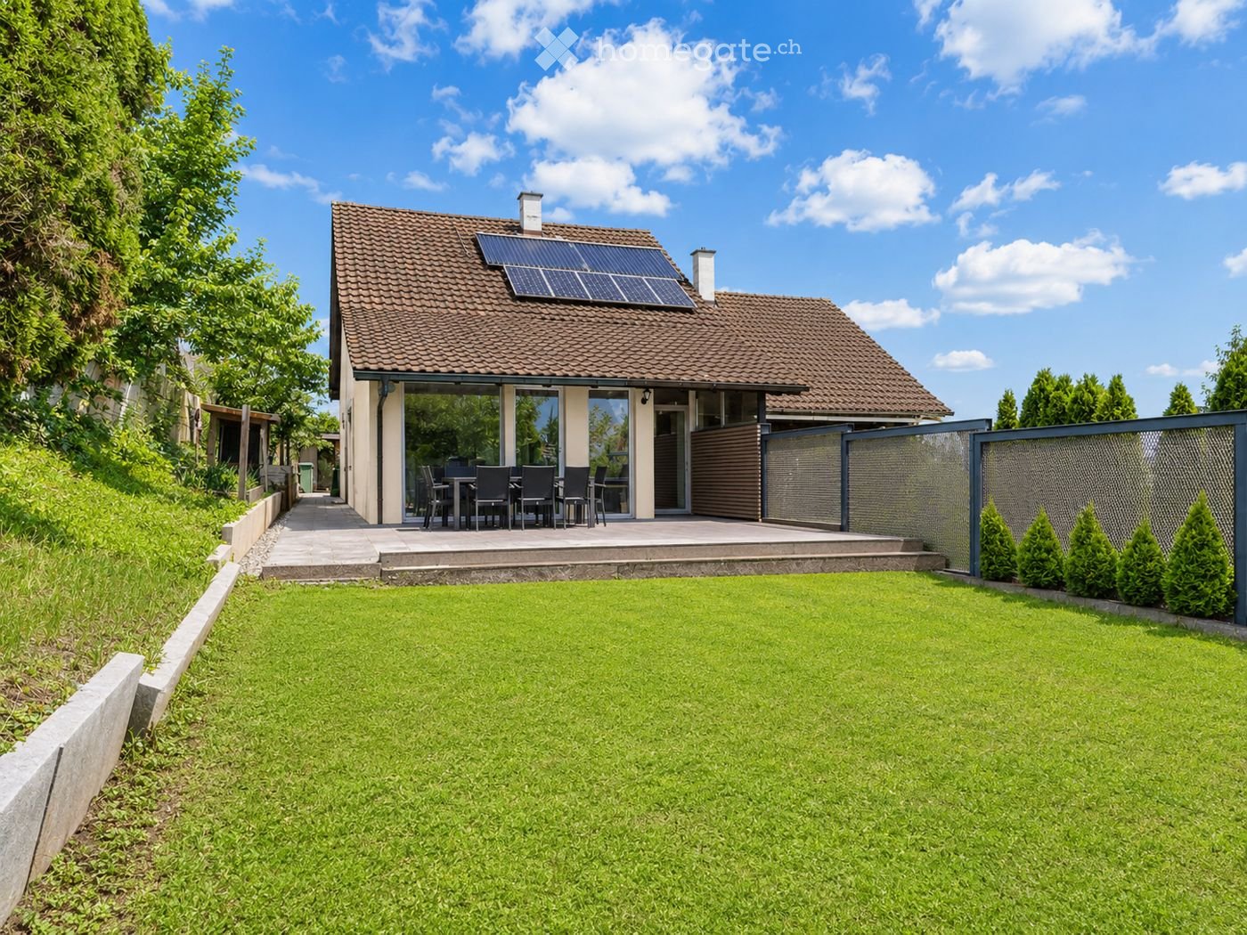Two-story house with a tiled roof, solar panels, and a modern design. The house features a raised ground floor, a patio with outdoor furniture, and a well-maintained lawn.