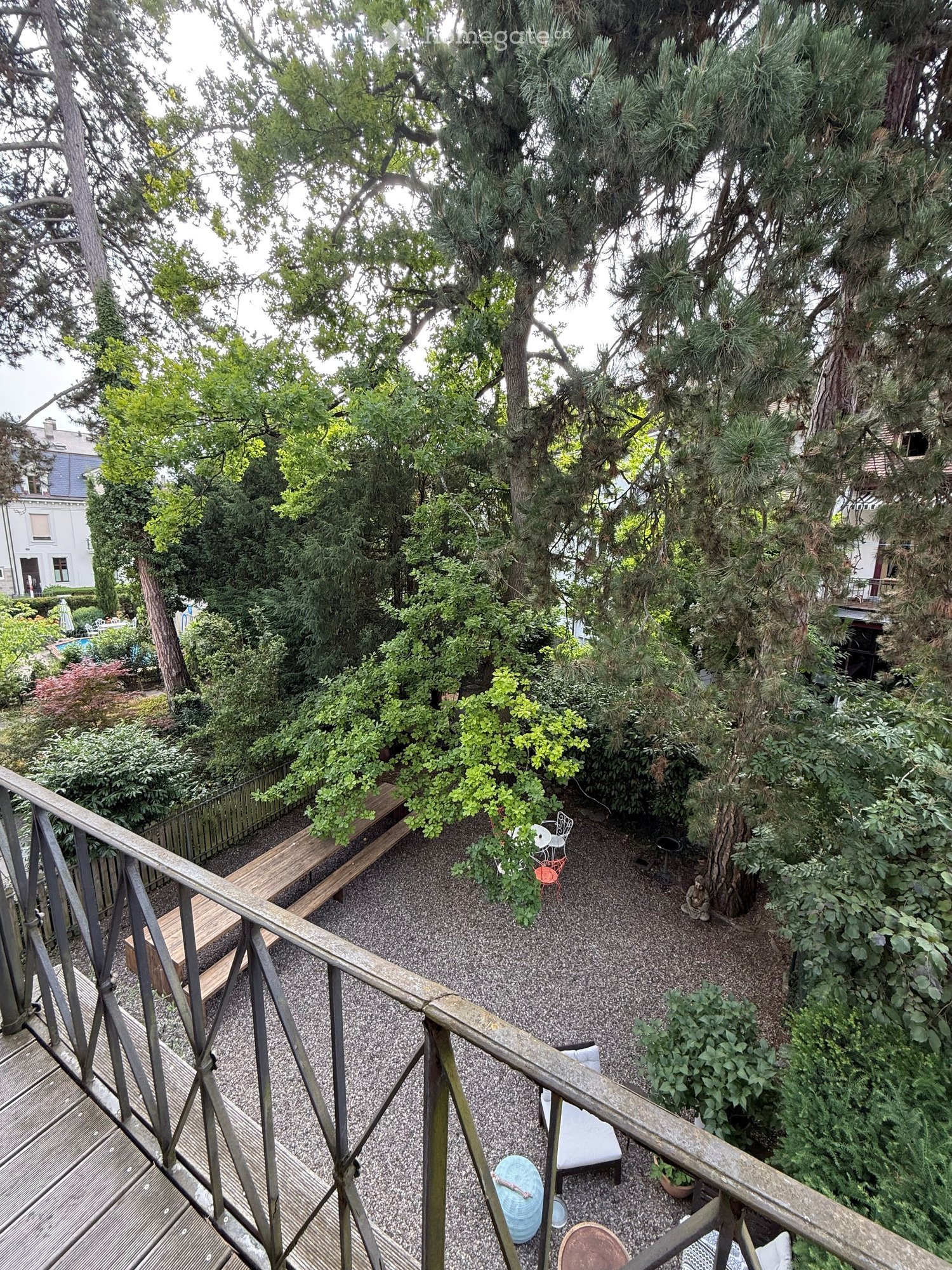 Garden area with gravel ground, wooden bench, small seating area with chairs and table, surrounded by trees and greenery.