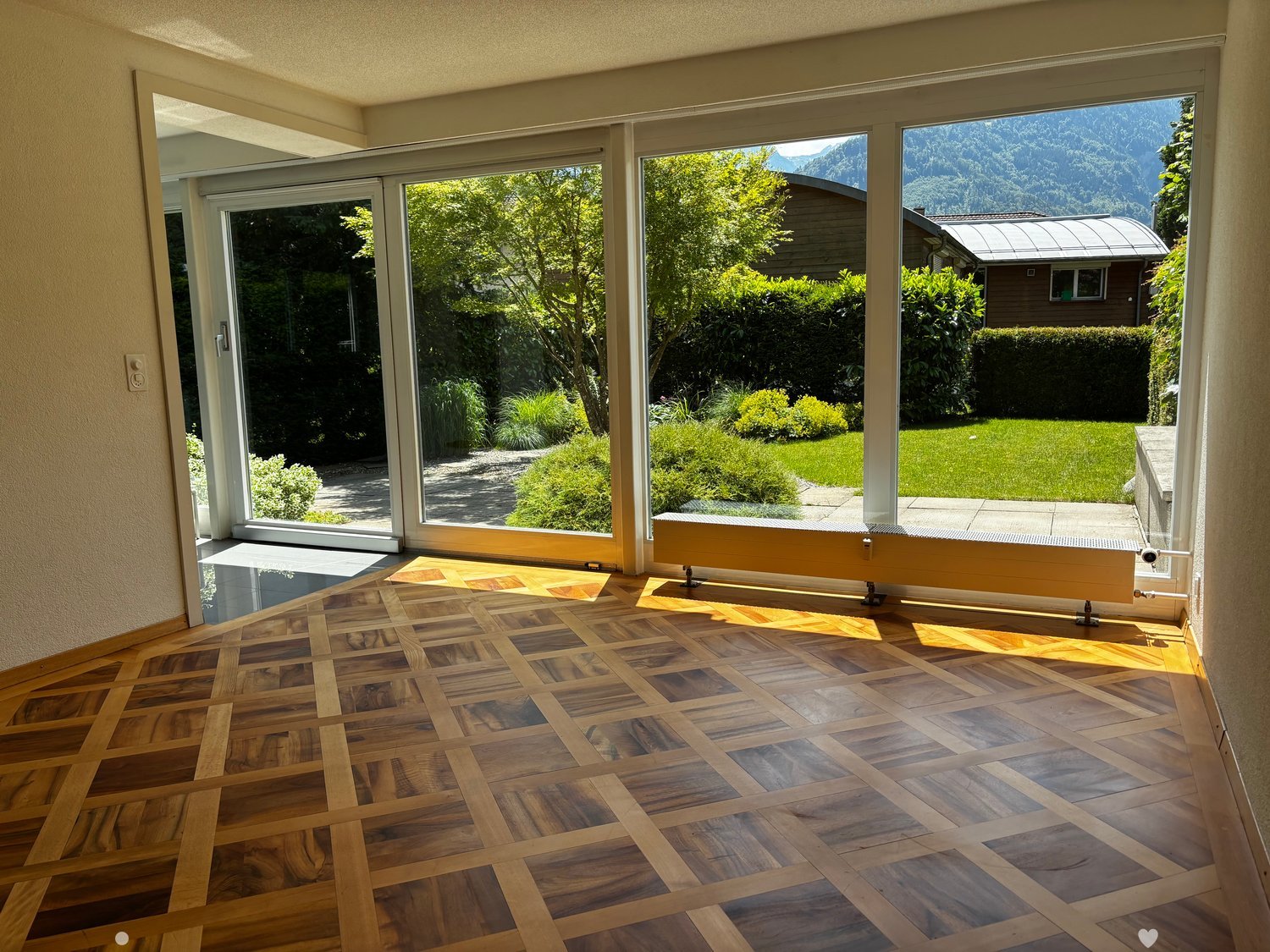 Empty room with wooden herringbone floor, large glass sliding doors opening to a garden, radiator, and a view of mountains and greenery.