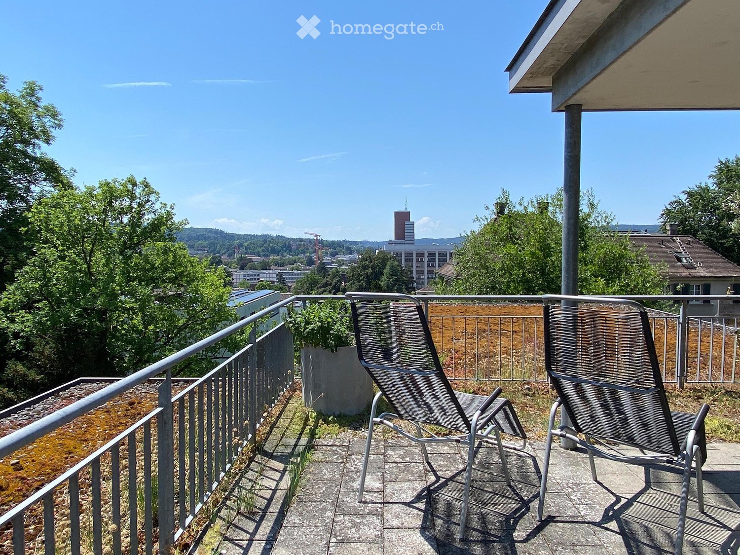 Balcony with two lounge chairs, metal railing, and a view of the city and surrounding greenery.