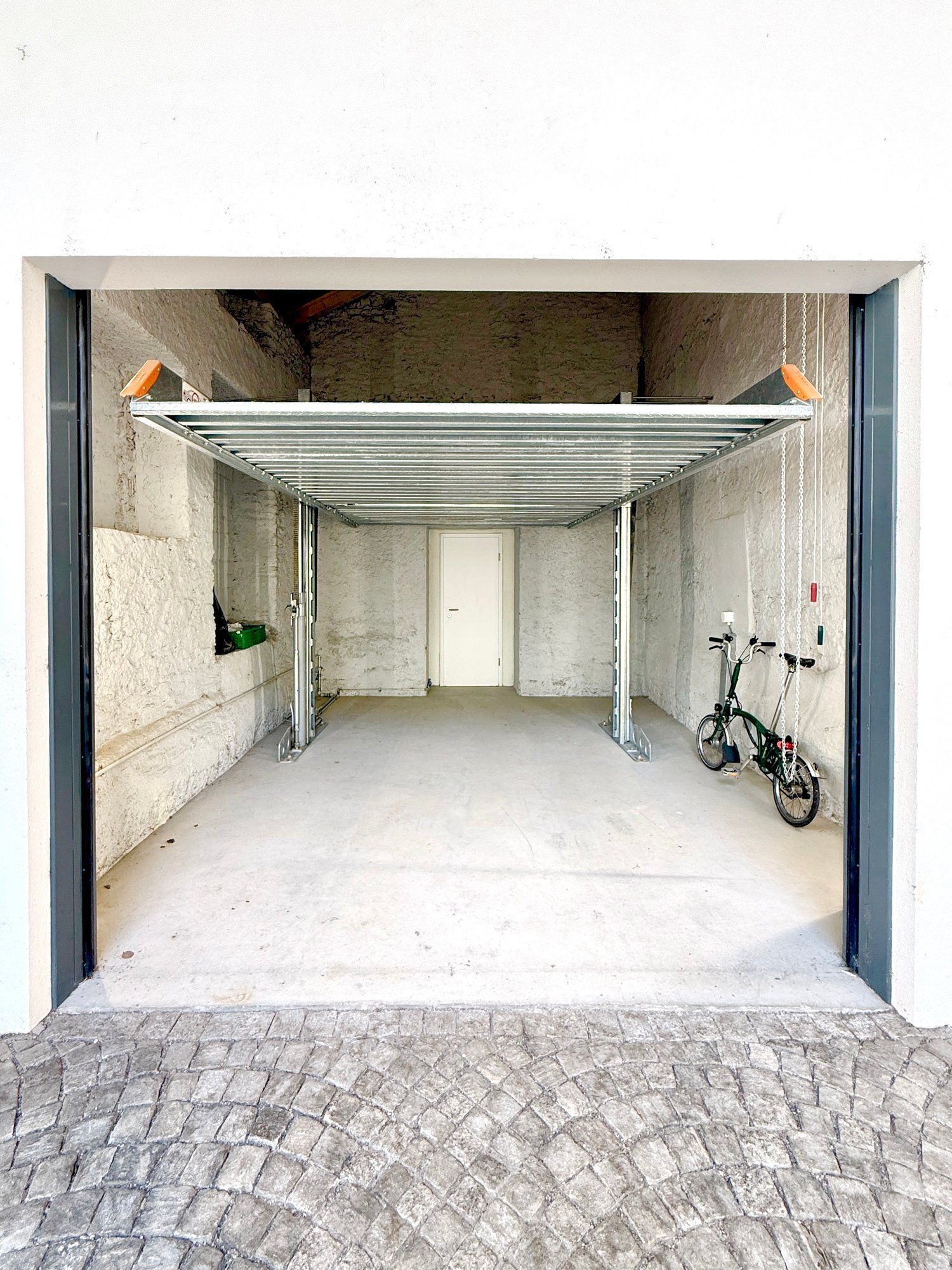 A covered, open-air corridor with concrete walls and a metal roof structure, featuring a bicycle parked on the right side and a white door at the far end.