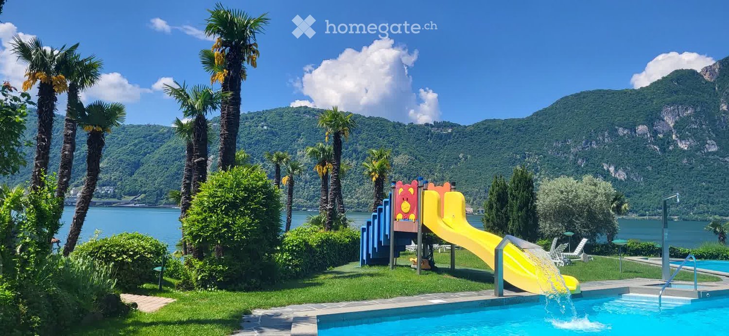 Outdoor area with a swimming pool, water slide, palm trees, and a scenic lake view with mountains in the background.