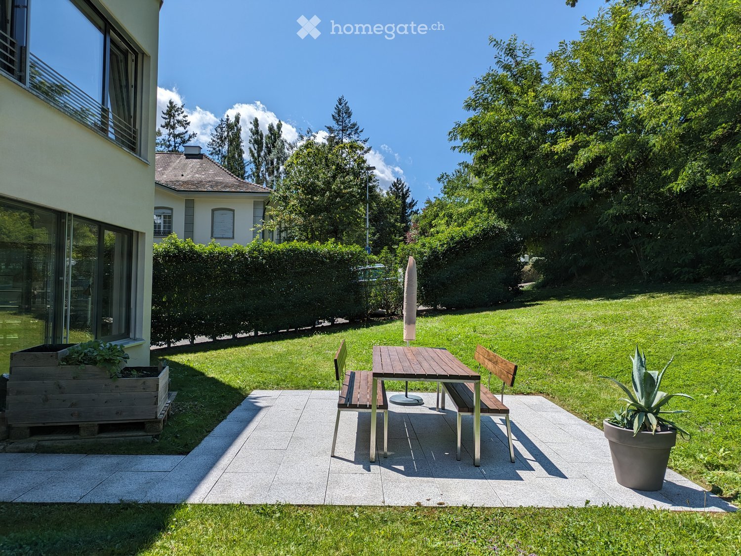 Garden with a wooden table, two benches, an umbrella, and a potted plant, surrounded by a well-maintained lawn and trees.