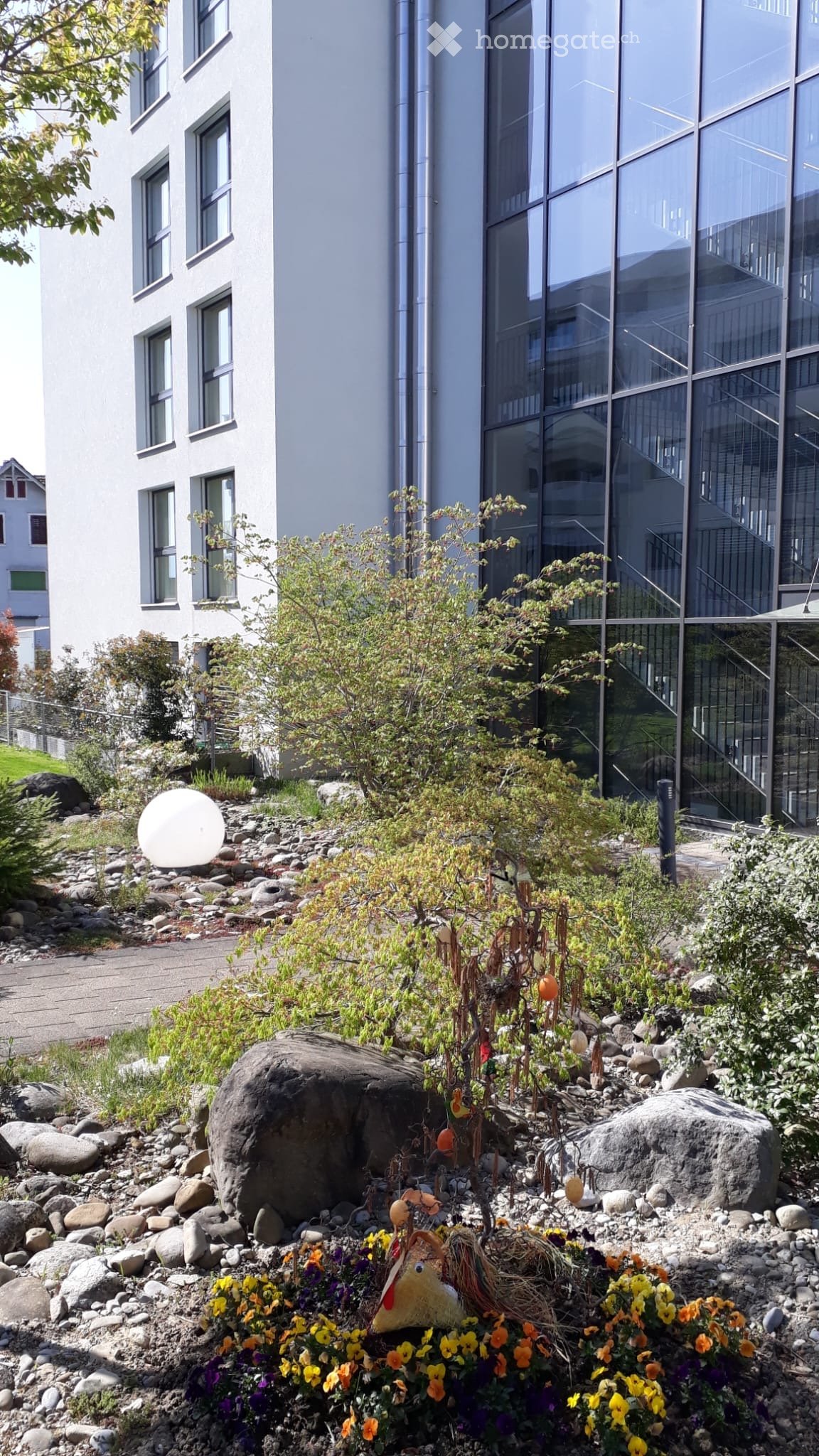 Garden area with rocks, plants, flowers, and a decorative turkey figure near a modern building with large windows and a staircase.