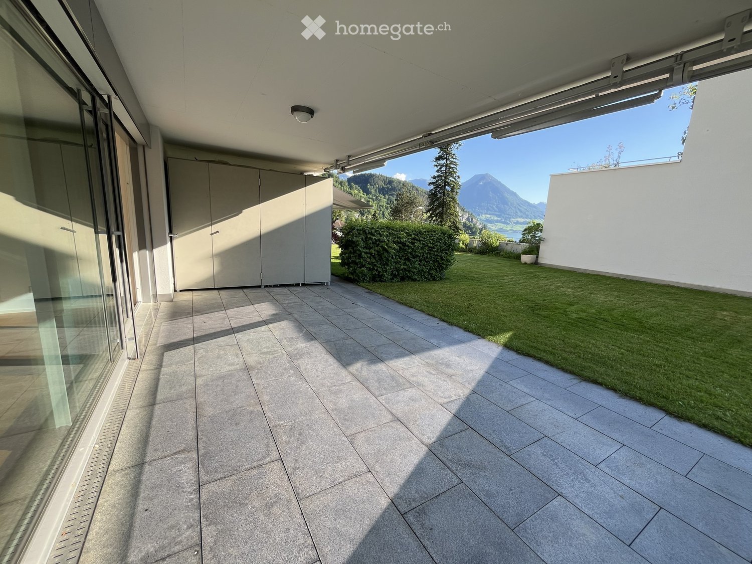 Modern balcony with tiled flooring, large glass sliding doors, and a view of a green lawn and mountains.
