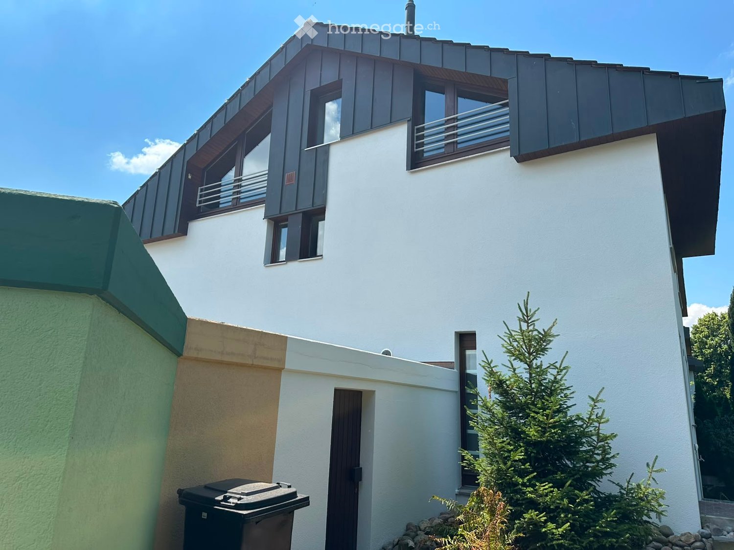 Modern two-story house with white walls, dark roof, and large windows. Features a balcony on the upper floor and a small garden area with a pine tree.