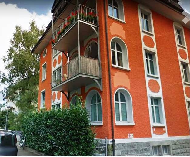 Apartment building with 3 stories, balcony on the first floor, brick and white exterior walls, large glass windows, small garden