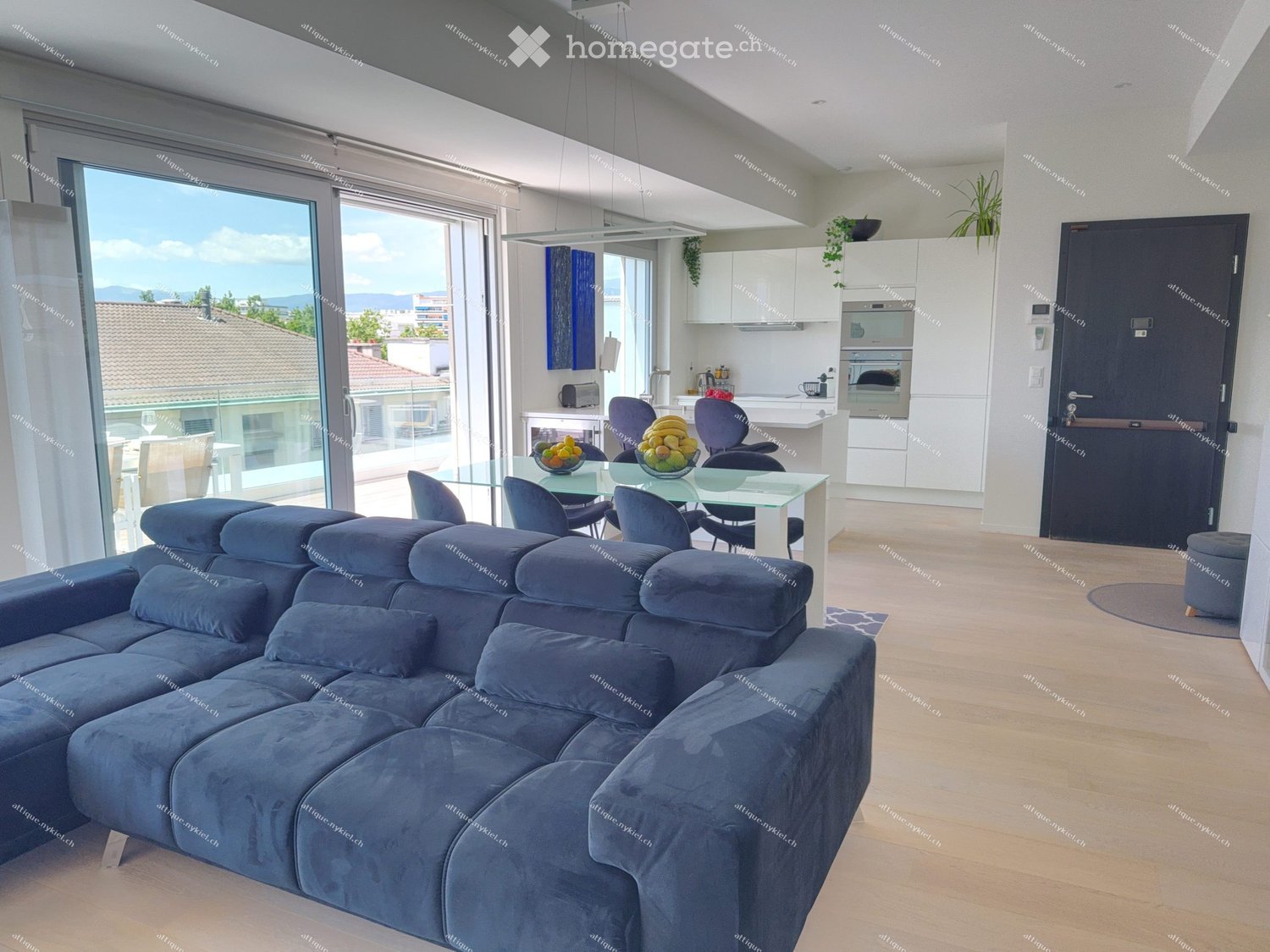 Modern living and dining area with a large sectional sofa, coffee table, dining table with chairs, and an open kitchen with white cabinets, an oven, and a refrigerator. The space features large windows with a view of the city and mountains.
