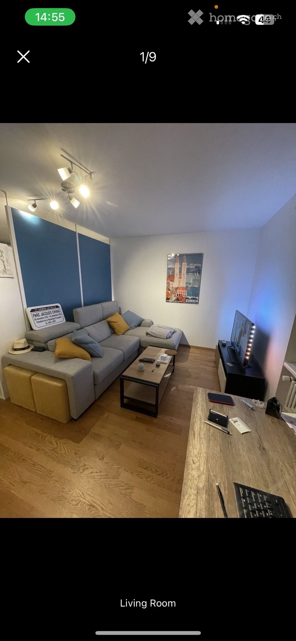 Modern living room with a grey sectional sofa, wooden coffee table, TV stand, and a small desk with a laptop and keyboard. The room features wooden flooring, a blue accent wall, and decorative artwork.