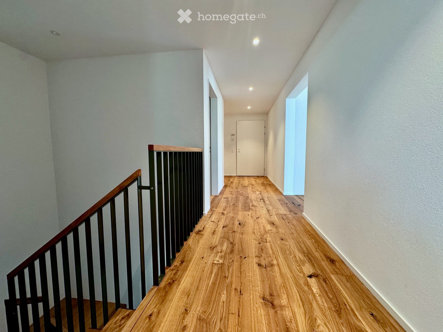 A modern hallway with wooden flooring, white walls, and a staircase with a wooden handrail and metal balustrade. The hallway features recessed lighting and a closed door at the end.