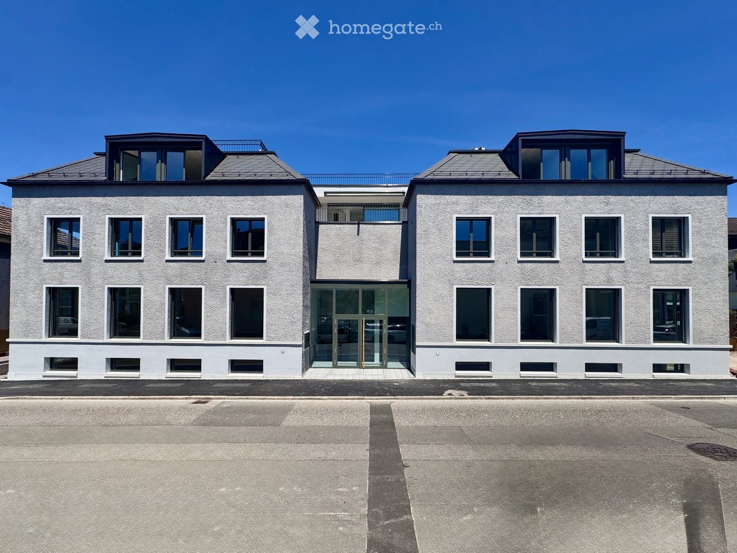 Modern two-story building with light grey facade, multiple windows, and a central glass entrance. The roof features small dormer windows and a small balcony area.