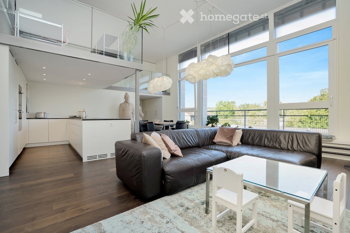 Modern open-concept living and dining area with large windows offering a view, a black leather sectional sofa, a glass coffee table with white chairs, a white kitchen island, and a dining table with chairs in the background.