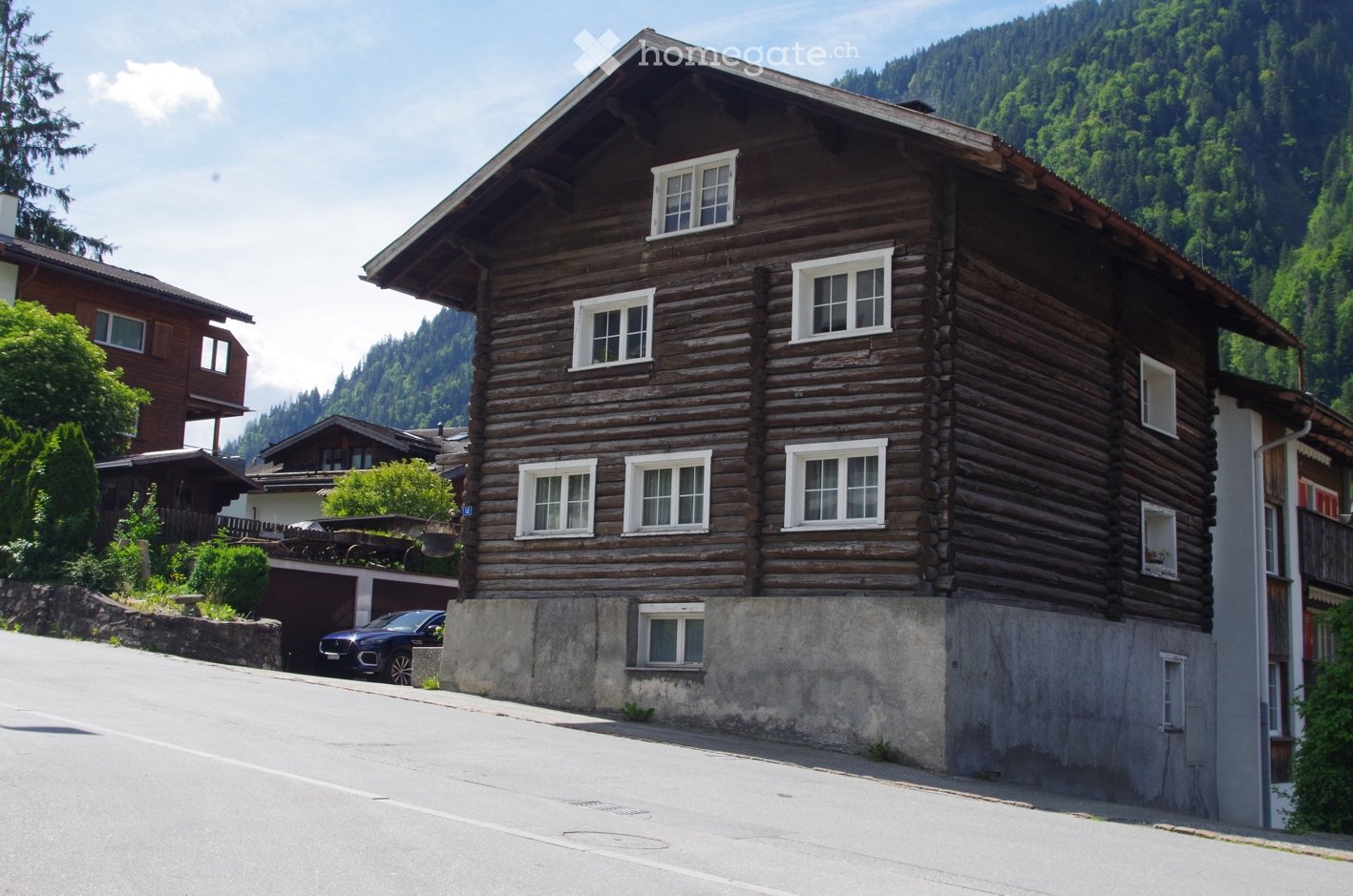 Traditional wooden house with multiple windows and a sloped roof, situated in a scenic mountainous area with neighboring houses and greenery.