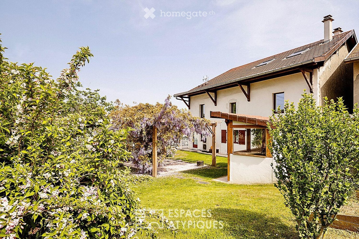 A two-story house with a tiled roof, white exterior, and a covered wooden porch. The house is surrounded by a well-maintained garden with lush greenery and flowering shrubs.