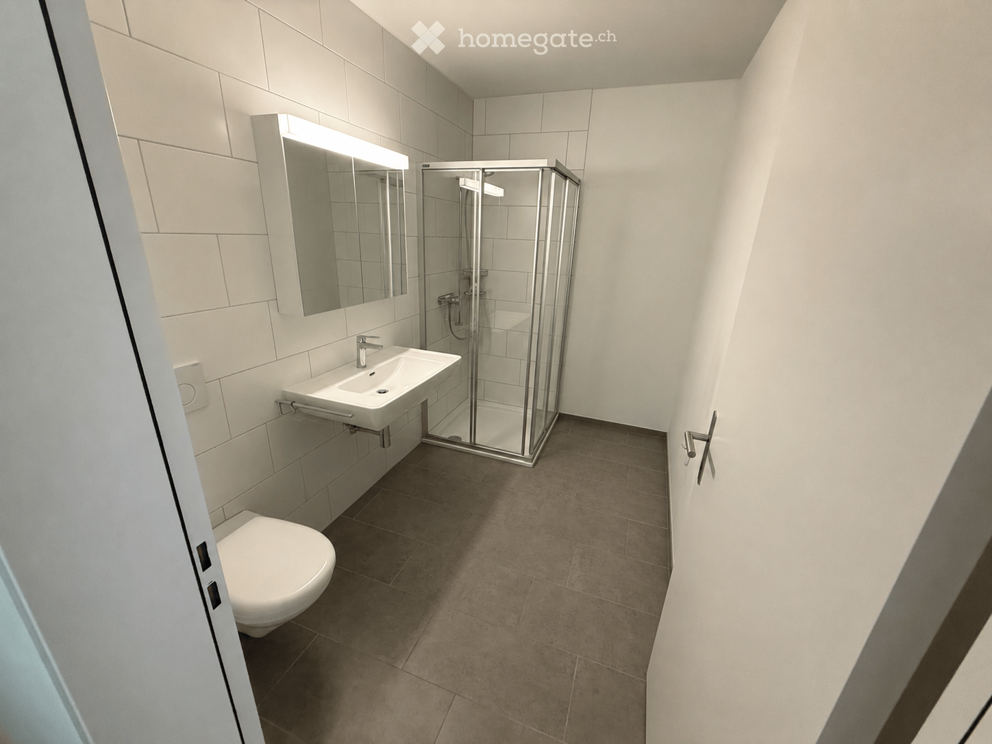 Modern bathroom with white tiles, a toilet, a sink with a mirror, and a glass-enclosed shower stall.
