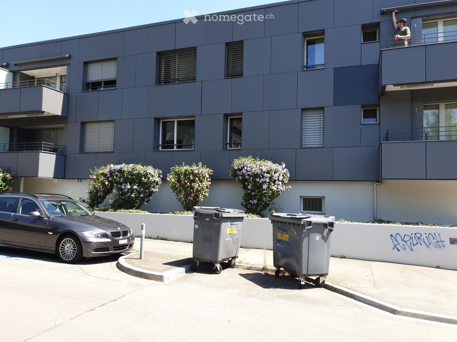 Modern multi-story building with grey facade, multiple windows, balconies, and small trees in front. A car is parked nearby, and two large trash bins are visible.