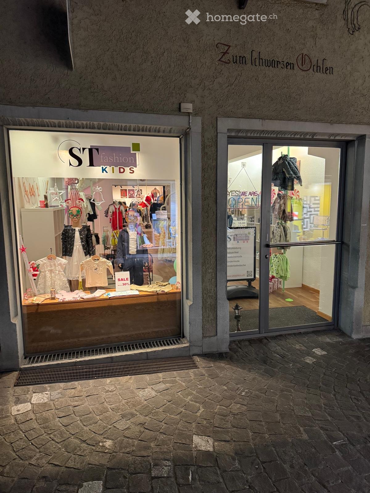 Storefront with glass doors, display of children's clothing, sign indicating sale, and a lamp outside