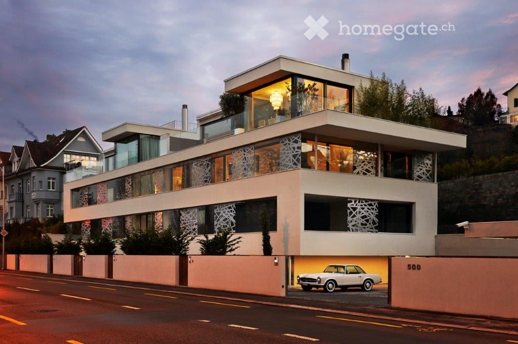 Modern two-story house with large glass windows, white exterior, and a sleek design. Features a raised ground floor and a parked vintage car in the driveway.