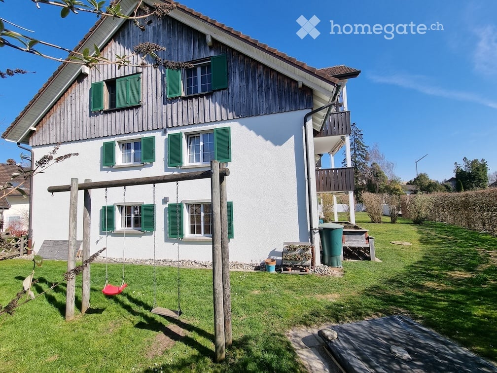 Two-story house with white walls, green shutters, and a wooden balcony. The front yard features a swing set and a small garden area.
