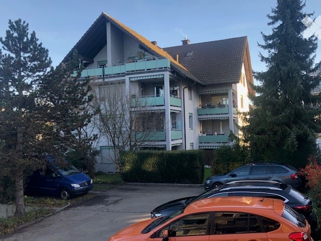 Multi-story residential building with balconies, surrounded by trees and parked cars.