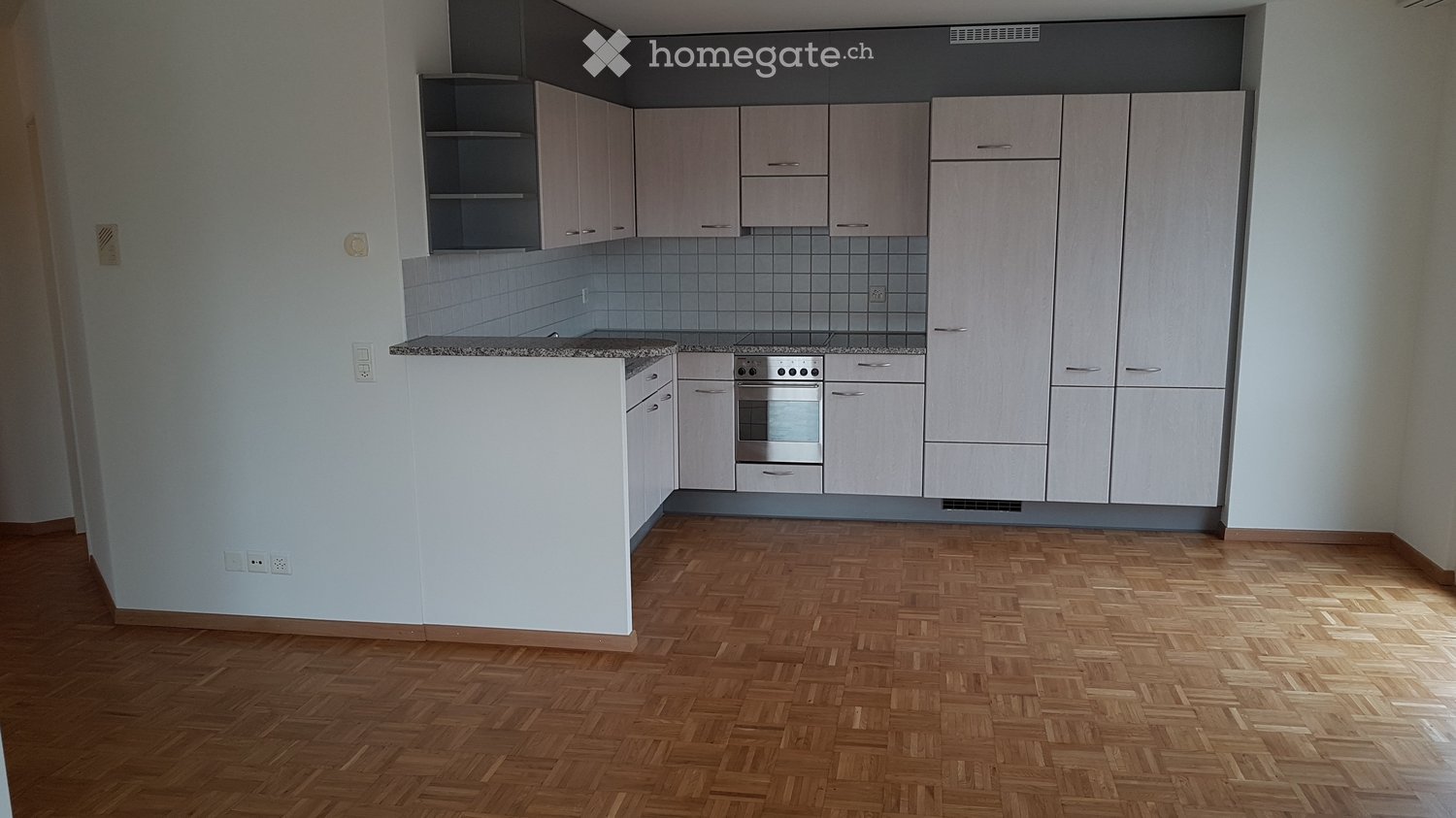 Modern kitchen with light wood cabinets, stainless steel oven, and granite countertop. The floor is made of herringbone-patterned wood.