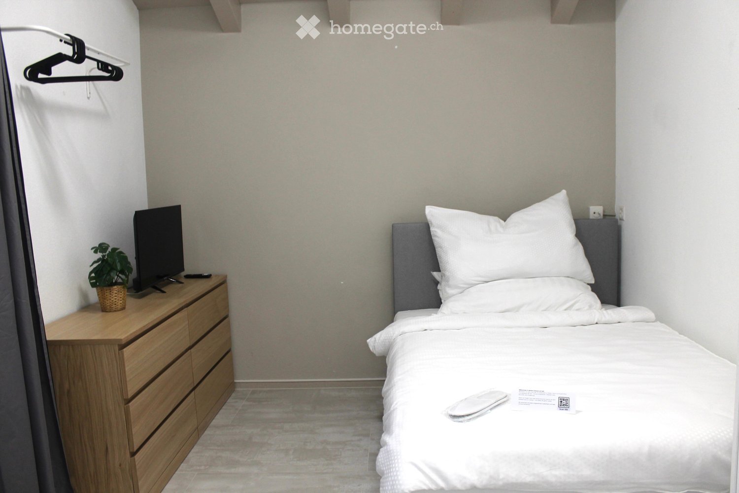 A modern, minimalist bedroom with a single bed, a wooden nightstand, a television, and a small potted plant. The room features light-colored walls and a tiled floor.